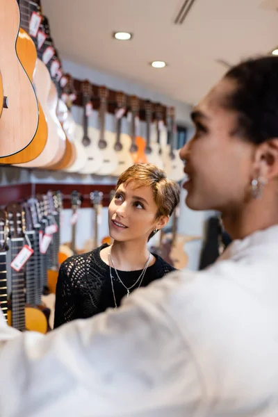 Усміхнений клієнт дивиться на акустичні гітари поблизу розмитого афроамериканського продавця в музичному магазині — стокове фото