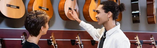 Positivo vendedor afroamericano apuntando a la guitarra acústica cerca del comprador en la tienda de música, bandera - foto de stock