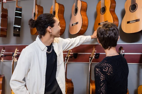 Vendedor afroamericano mirando guitarras acústicas cerca del cliente en la tienda de música - foto de stock