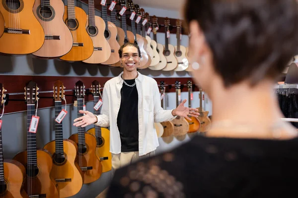 Sonriente vendedor afroamericano señalando con las manos cerca de las guitarras acústicas y el cliente borroso en la tienda de música - foto de stock