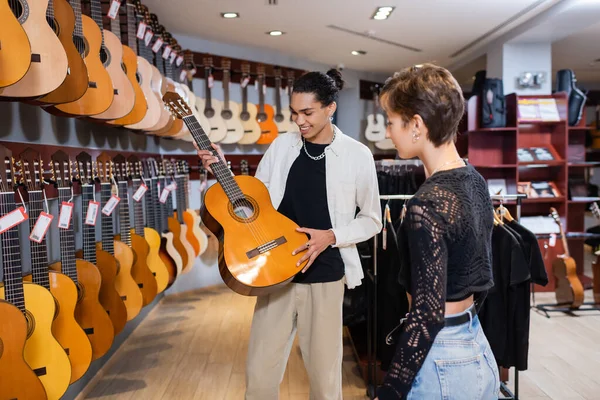 Venditore africano americano sorridente che tiene la chitarra acustica vicino all'acquirente nel negozio di musica — Foto stock