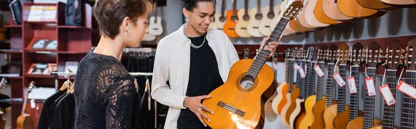 Позитивний афроамериканський продавець тримає акустичну гітару поблизу жінки в музичному супроводі, банер — стокове фото