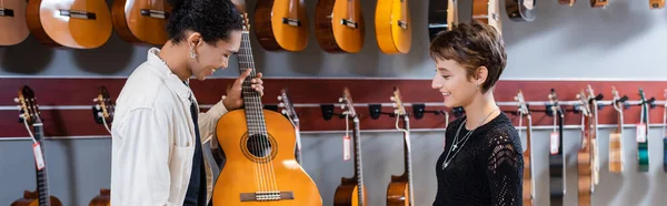 Vista lateral del vendedor afroamericano sosteniendo la guitarra acústica cerca del cliente en la tienda de música, bandera - foto de stock