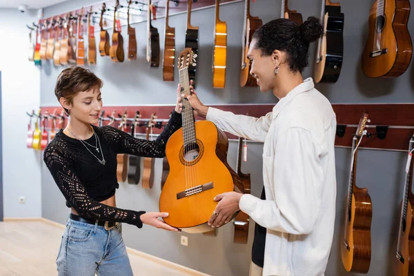 Vendedor afroamericano dando guitarra acústica a cliente sonriente en tienda de música - foto de stock