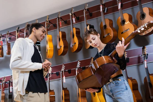 Vista a basso angolo di sorridente venditore afroamericano guardando donna con chitarra acustica nel negozio di musica — Foto stock