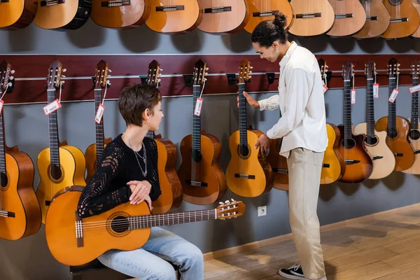 Vista laterale del venditore africano americano sorridente che prende la chitarra acustica vicino al cliente nel negozio di musica — Foto stock