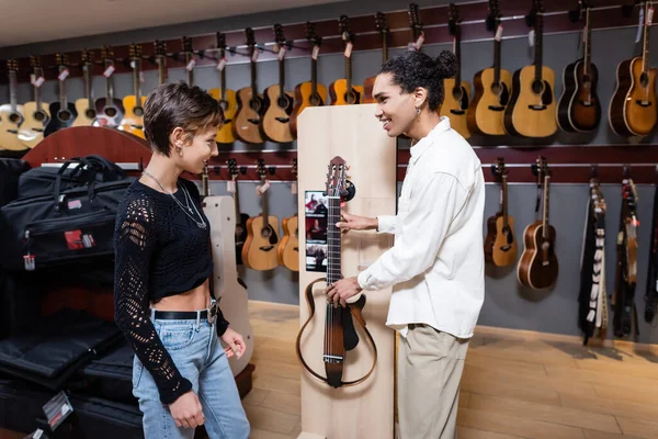 KYIV, UKRAINE - FEBRUARY 16, 2022: Side view of african America seller holding electronic Guitar near customer in music store — стокове фото