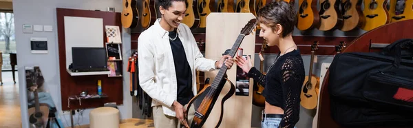 KYIV, UKRAINE - FEBRUARY 16, 2022: Smiling buyer looking at electric guitar near african american seller in music shop, banner — Stock Photo