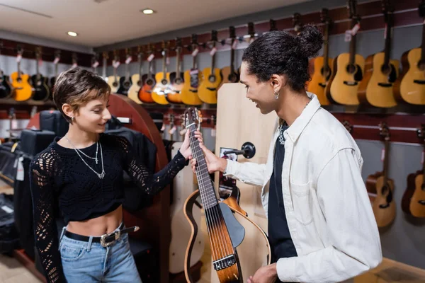 KYIV, UCRANIA - 16 DE FEBRERO DE 2022: Alegre vendedor afroamericano sosteniendo la guitarra eléctrica cerca de una joven en la tienda de música - foto de stock