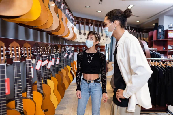 Young woman in medical mask looking at acoustic guitars near african american seller in music store — Foto stock