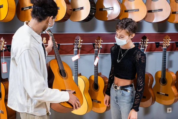 Продавец африканской Америки держит акустическую гитару рядом с клиентом в медицинской маске в музыкальном магазине — стоковое фото