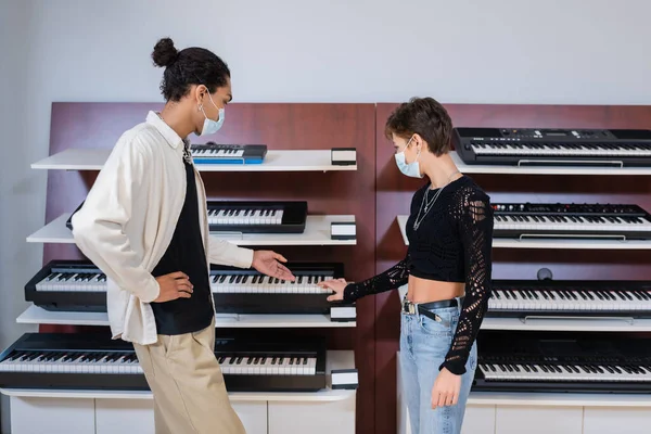 African american seller in medical mask pointing at synthesizer near customer in music store — Stock Photo