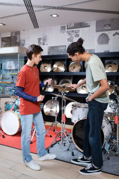 Side view of seller holding drumsticks near african american customer and drums in music store — Stock Photo