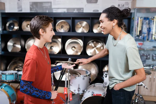 Side view of cheerful seller holding drumsticks near african american customer and drum set in store — Stock Photo