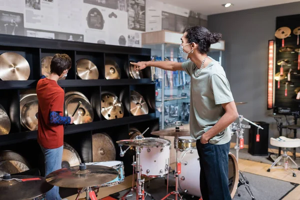 Client afro-américain en masque médical pointant vers la plaque de tambour près du vendeur dans un magasin de musique — Photo de stock