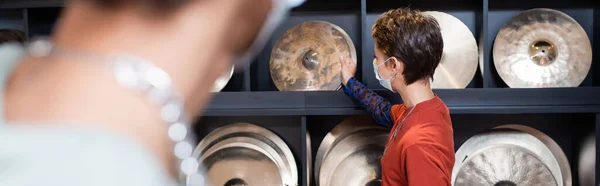 Seller in medical mask taking drum plate near african american buyer in music store, banner — Stock Photo