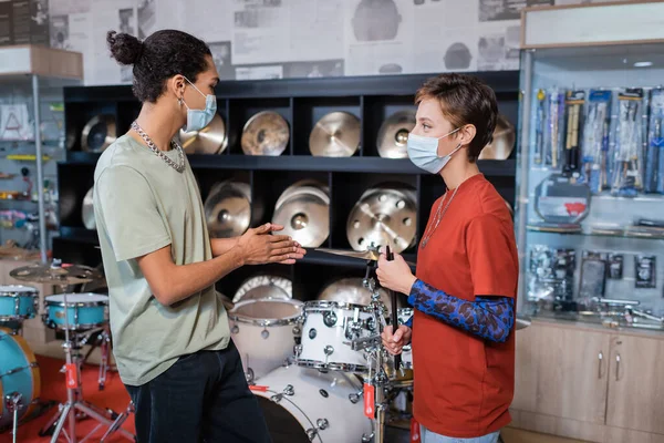 Side view of seller holding drumsticks near african american customer in medical mask in music store — Fotografia de Stock