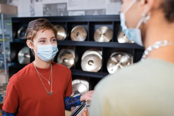 Vendedor em máscara médica conversando com cliente afro-americano desfocado na loja de música — Fotografia de Stock