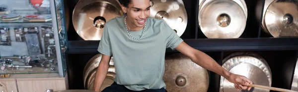 Cheerful african american customer holding drumstick near plates in music store, banner — Stock Photo