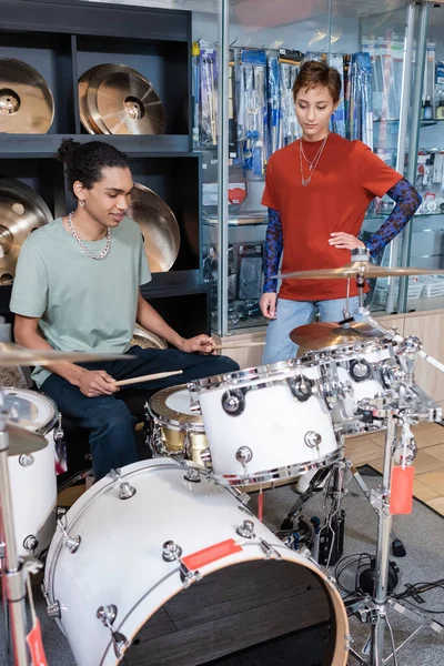 Vendedor de pie cerca de cliente afroamericano tocando la batería en la tienda de música - foto de stock