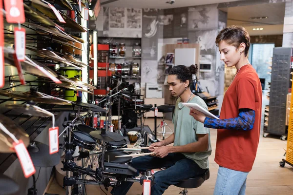 Vue latérale du vendeur tenant une tablette numérique près d'un client afro-américain jouant de la batterie électrique en magasin — Photo de stock