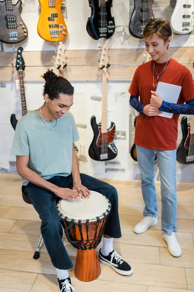 KYIV, UKRAINE - 16 FÉVRIER 2022 : Vendeur souriant tenant une tablette numérique près d'un client afro-américain jouant au djembé dans un magasin de musique — Photo de stock