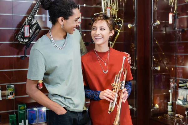 Mulher alegre segurando trompete e olhando para o namorado afro-americano na loja de música — Fotografia de Stock