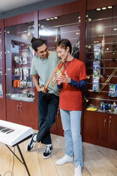 Cheerful woman holding trumpet near african american boyfriend and synthesizer in music store — Stock Photo