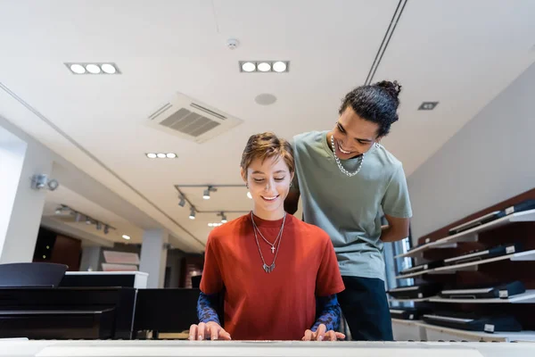 Smiling woman playing synthesizer near african american boyfriend in music store — Stockfoto