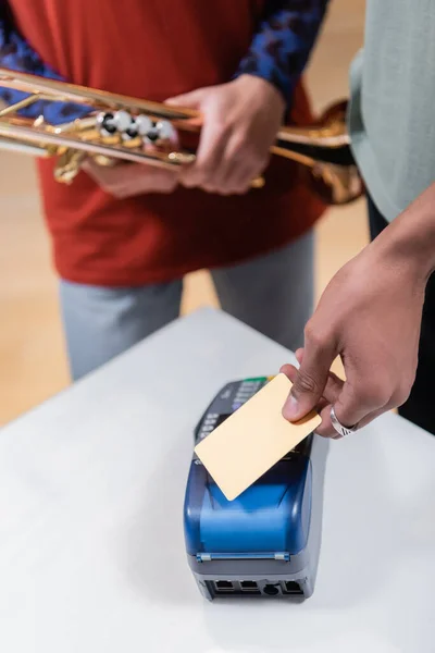 Vista cortada do homem americano africano que paga com cartão de crédito perto da namorada com trompete na loja de música — Fotografia de Stock