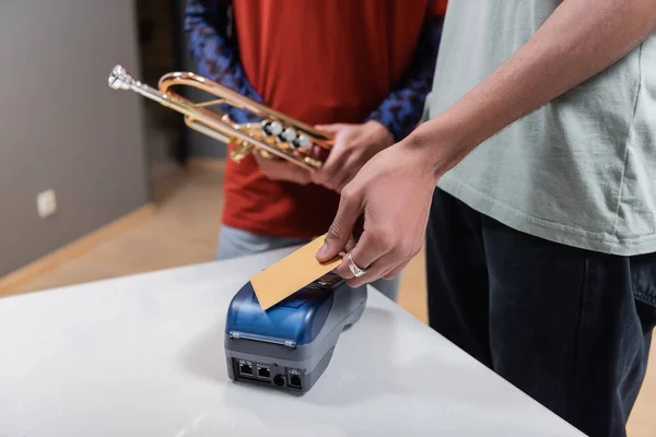 Cropped view of african american man paying with credit card near blurred girlfriend with trumpet in music store — Stock Photo