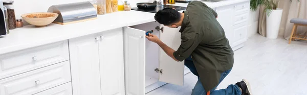Young muslim man with screwdriver fixing cabinet under worktop in kitchen, banner — Foto stock