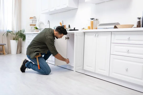 Side view of muslim man with tool belt fixing kitchen cabinet — Fotografia de Stock
