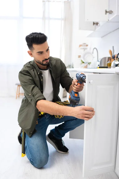 Muslim man with electric screwdriver fixing door of kitchen cabinet — Stock Photo