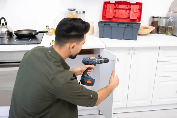 Young arabian man with electric screwdriver fixing door of kitchen cabinet near toolbox on worktop — Foto stock