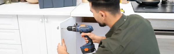 Muslim man with electric screwdriver fixing cupboard in kitchen, banner — Stock Photo