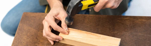 Cropped view of man holding hammer and nail on wooden board at home, banner — Stockfoto