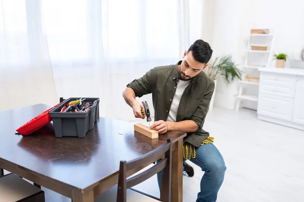 Arabian carpenter holding hammer near nail on wooden board and toolbox at home — Stockfoto