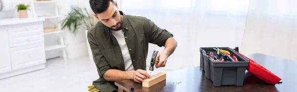 Arabian man holding hammer near nail on board near tools on table at home, banner — Fotografia de Stock