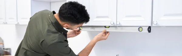 Arabian man measuring kitchen cupboard with spirit level at home, banner — Stock Photo
