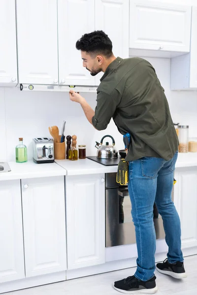 Side view of arabian man with spirit level measuring kitchen cupboard at home — Fotografia de Stock