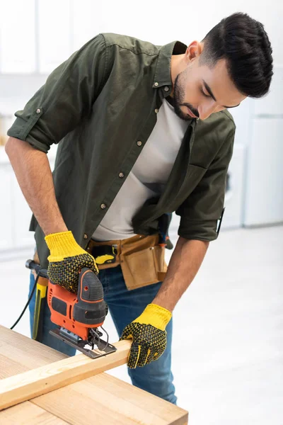 Arabian man with tool belt using jigsaw machine on wooden plank at home — Fotografia de Stock