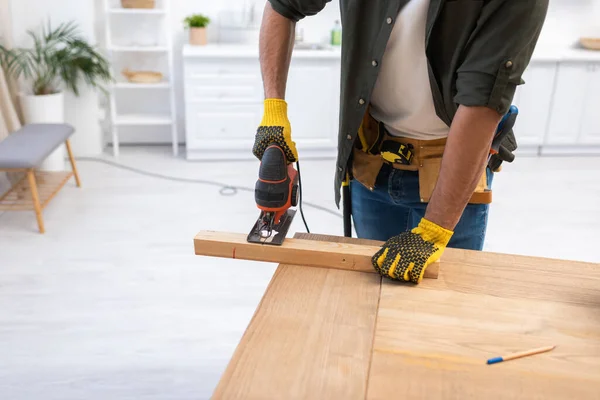 Cropped view of man in gloves holding jigsaw machine while sawing plank at home — Photo de stock