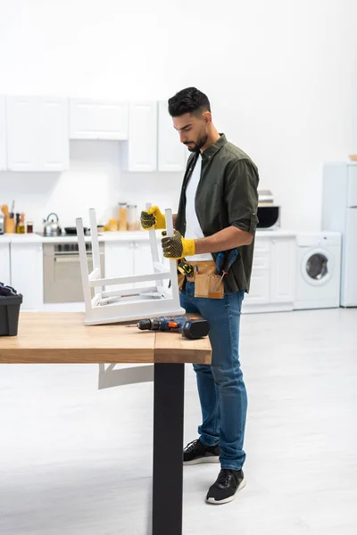 Young muslim craftsman in gloves holding chair near tools at home — Stock Photo