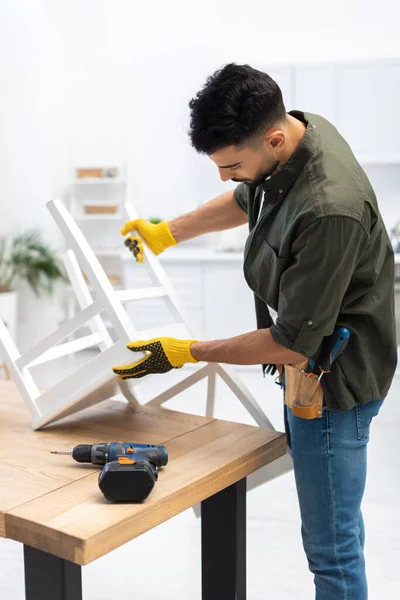 Arabian craftsman in gloves holding chair near electric drill at home — Fotografia de Stock