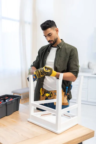 Arabian man in gloves holding electric drill near chair on table at home — Fotografia de Stock