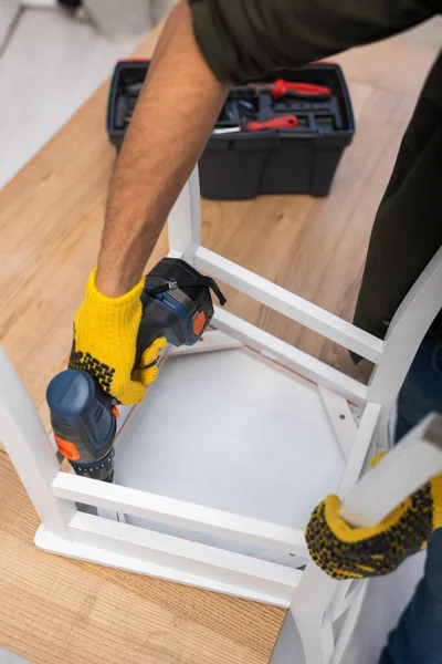 Cropped view of man in gloves repairing chair with electric screwdriver near blurred toolbox at home - foto de stock