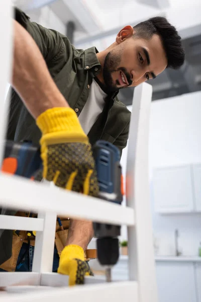 Smiling muslim craftsman fixing chair with blurred electric screwdriver at home — Stock Photo