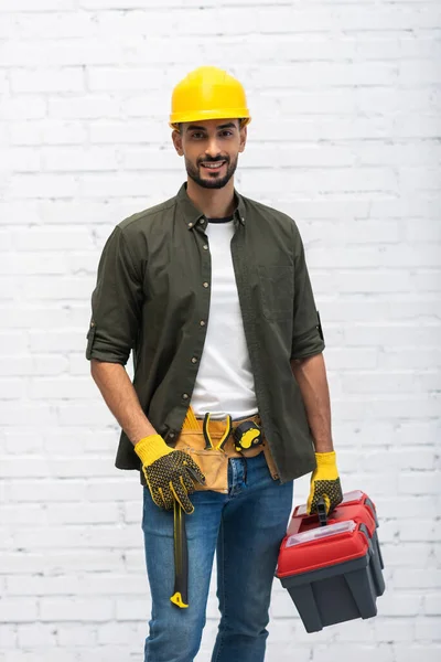 Positive muslim man in hardhat holding toolbox at home - foto de stock