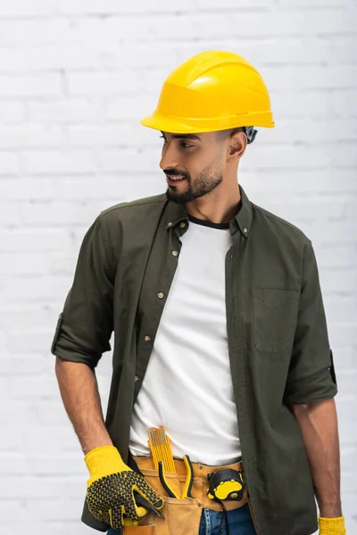 Smiling muslim man in hardhat and gloves touching tool belt at home — Stock Photo